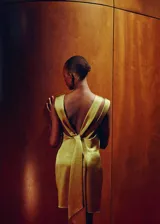 Vestido corto de satén color amarillo, con diseño sin mangas, cuello redondo y detalle de fruncido y nudo en el hombro. Presenta un corte recto y espalda abierta.