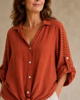 Camisa de lino color terracota, de corte holgado, con cuello camisero, cierre frontal de botones y mangas largas con detalles perforados y trabilla para arremangar.