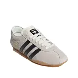 Championes Adidas modelo Tokyo de corte bajo, confeccionados en gamuza color beige con las icónicas tres tiras laterales en cuero negro. Presenta suela de caucho color caramelo y detalle de logo dorado en el lateral.