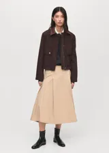 Chaqueta corta de algodón en color marrón oscuro, con cuello de efecto piel en tono contrastante. Presenta cierre frontal con cremallera y botones a presión, además de dos bolsillos frontales con solapa y cierre de botón.