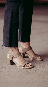 Sandalia de cuero color taupe con taco cuadrado de 6 cm, tira frontal ancha y tira en el tobillo con detalle metálico dorado y hebilla circular.