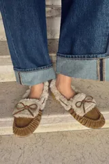 Mocasín plano tipo náutico confeccionado en serraje color tostado, con interior y borde superior forrados en tejido efecto borreguito. Presenta detalle fruncido en la puntera redonda y borlas decorativas en la parte frontal.
