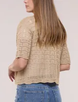 Bolero corto de punto beige, con diseño abierto sin botones y mangas cortas. Presenta un patrón de tejido calado con detalles geométricos y terminaciones onduladas en los bordes.