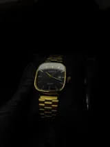 Reloj Casio con caja cuadrada de bordes redondeados en color dorado y esfera negra. Cuenta con malla de acero inoxidable dorada, manecillas e índices dorados, y ventana de fecha en la posición de las tres.