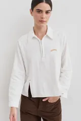 Remera de manga larga color blanco con cuello polo y cierre frontal parcial. Presenta costuras a la vista en contraste y un pequeño bordado en el pecho. El diseño incluye una abertura lateral en el ruedo.