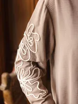 Camisa de color beige con cuello clásico y cierre frontal de botones. Presenta un diseño de bordado lineal en color blanco que recorre las mangas largas.