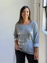 Remera gris de manga larga con cuello redondo y diseño holgado. Presenta un aplique frontal rectangular con detalles de lentejuelas plateadas y estampado tipográfico.