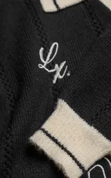 Sweater de punto negro con cuello polo en color crema, manga larga y diseño oversize. Presenta bordados en el frente con la inscripción 'Ravers', el número '90' y letras cursivas, con terminaciones en punto rib en puños y cuello.