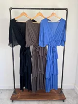 Vestido midi de seda italiana color azul, con escote en V, mangas murciélago amplias, falda de tres volados y cintura elastizada con lazo ajustable al tono y hebilla circular dorada.
