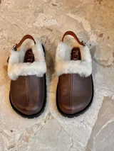 Zueco de cuero color marrón con interior forrado en piel sintética blanca, tira trasera ajustable con hebilla metálica y suela de goma negra con relieve.