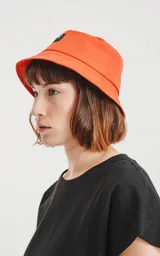 Gorro tipo bucket de tela color naranja vibrante, con un pequeño parche negro en el frente que tiene un logo bordado en forma de tijeras cruzadas.