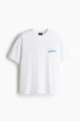 Remera de algodón blanca con cuello redondo acanalado y hombros caídos. Presenta un pequeño estampado gráfico en azul en el pecho con la palabra Sevilla.