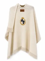 Ruana marfil de tejido acanalado con flecos en los bordes, parche de cuerina con logo de la marca en el pecho y un escudo bordado con detalles en dorado y azul.