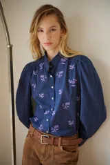 Camisa azul marino con estampado floral en color lila, cuello mao con volantes, cierre frontal de botones y mangas largas abullonadas con puños elásticos.