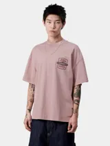 Camiseta de manga corta Carhartt WIP, color rosa pálido, con cuello redondo y corte holgado. Presenta un pequeño parche estampado en el pecho con texto que incluye "Detroit's Extra Rugged" y el logo de Carhartt.