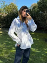 Camisa blanca de silueta entallada con alforzas frontales, cuello clásico, cierre de botones y mangas largas.
