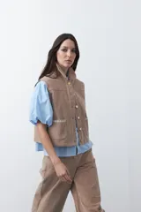 Chaleco de pana color marrón oscuro, con cuello camisero, cierre frontal de botones metálicos dorados y dos bolsillos frontales con detalle de bordado geométrico.