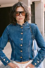 Chaqueta de denim azul con cuello redondo, manga larga y cierre frontal con botones dorados. Presenta costuras verticales decorativas y puños con botones a juego.