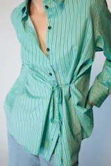 Camisa de manga larga con cuello solapa, cierre frontal con botones y diseño cruzado con lazada lateral en el mismo tejido. Presenta un estampado de rayas verticales en tonos verdes y azules.