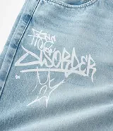 Pantalón de jean corte baggy, color celeste claro con efecto desgastado y estampado de lettering estilo grafiti en color blanco. Presenta cintura con trabillas, cierre frontal con botón y cremallera, y bolsillos laterales y traseros.