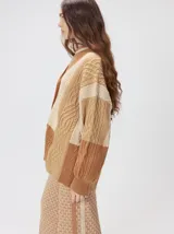 Cardigan oversize de tejido de punto grueso con diseño de bloques de color en tonos beige y marrón. Presenta cuello en V, cierre frontal con botones, mangas largas abullonadas y parches decorativos en el pecho y el hombro.