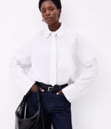 Camisa maxi oversized blanca con finas rayas verticales negras, de manga larga con puños anchos, cuello camisero con corbata incorporada, bolsillo en el pecho y cierre frontal con botones ocultos.