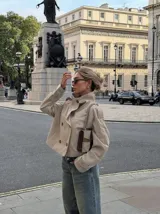 Chaqueta corta tipo trench color beige, con doble botonadura frontal, cuello alto y mangas largas con puños ajustables. Presenta un diseño estructurado con hombreras y solapas.