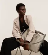 Bolso grande tipo hobo desestructurado, color gris claro con acabado ligeramente brillante o envejecido. Cuenta con dos asas cortas y una correa larga desmontable. Presenta detalles de cierres o tiradores decorativos en los laterales y cierre principal superior con cremallera.