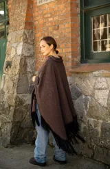 Poncho de tejido suave color marrón chocolate, con diseño amplio y relajado. Presenta detalles artesanales de costura en los bordes y en el bolsillo frontal, además de flecos largos en color negro en todo el ruedo.