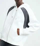 Saco con capucha y cierre frontal de cremallera, confeccionado en tejido de algodón y poliéster. Presenta un diseño bicolor con cuerpo blanco y paneles laterales en gris oscuro, mangas largas y bolsillos delanteros.