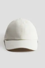 Gorra blanca de lino con ojales bordados, visera curvada y trabilla ajustable con cierre metálico en la parte posterior.