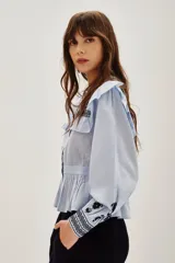 Camisa de algodón celeste con detalles de volados en los hombros y bordados en color negro en el pecho y las mangas. Presenta cierre frontal con botones y puños ajustados.