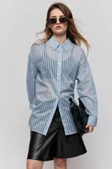 Camisa de rayas celestes y blancas de calce relajado, confeccionada en tejido liviano y semitransparente. Presenta cuello clásico, cierre frontal con botones, mangas largas con hombros caídos y aberturas laterales.