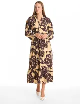 Vestido maxi de crepe con estampado floral en tonos beige y marrón oscuro. Presenta escote en V con botones frontales, mangas largas amplias con puños abotonados y cintura ajustable mediante cordón trenzado. La falda cuenta con un diseño de vuelos escalonados que aporta movimiento.