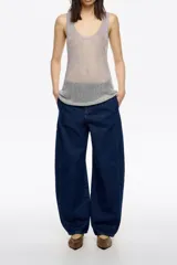 Pantalón de denim azul oscuro, corte barrel (tobillero con volumen en la cadera y estrechamiento hacia el tobillo). Presenta tiro medio, cierre frontal con corchete oculto y cremallera, y tiras ajustables con hebilla en la espalda.