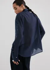 Camisa azul marino de lyocell y poliéster, con cuello camisero, manga larga, cierre frontal oculto con tapeta y detalles de apliques de strass y cristal en la parte delantera y las mangas.