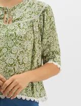 Blusa de gasa estampada con motivos botánicos en color verde, con escote redondo, abertura frontal con lazos de ajuste y mangas cortas. Presenta detalles de encaje en el escote, mangas y ruedo.