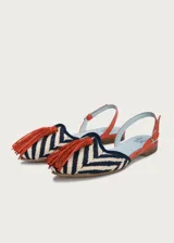 Sandalias planas destalonadas confeccionadas en rafia trenzada con patrón de zigzag en tonos azul marino y crudo. Tienen punta cerrada con una borla decorativa de flecos naranjas en el empeine, y una tira trasera ajustable de cuero naranja con hebilla metálica dorada.