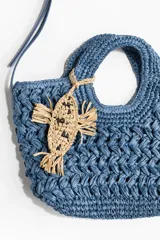 Cartera cruzada en paja trenzada de papel azul con doble asa, cierre magnético y bandolera ajustable, decorada con un dije colgante en forma de pez color beige.