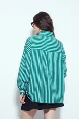 Camisa de silueta amplia y caída fluida, color celeste con rayas verticales blancas. Presenta cuello camisero clásico, cierre frontal con botones y un bolsillo aplicado en el pecho. Las costuras están resaltadas en azul oscuro.