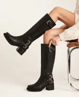 Bota de caña alta estilo biker, confeccionada en cuero negro. Presenta tres hebillas metálicas decorativas, cierre lateral y taco de madera.