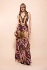 Vestido largo marrón de estilo boho con estampado paisley y floral en tonos tierra, rosado y celeste. Presenta escote en V profundo decorado con flecos dorados, pedrería y cordones entrecruzados, además de detalles de flecos en la zona de la cadera.