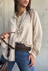 Camisa de corte holgado en color beige, con cuello clásico, cierre frontal de botones y mangas largas que presentan un delicado bordado tonal en los antebrazos.