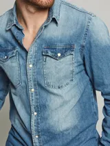 Camisa de jean de manga larga con cierre frontal de botones a presión, dos bolsillos frontales con solapa y puños abotonados.