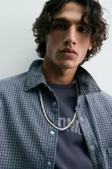 Camisa de manga larga con cuello solapa, bolsillo de plastrón en el pecho y cierre frontal con botones a presión. Presenta un patrón de cuadros vichy en tonos de azul desgastado.