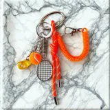 Llavero con argolla metálica plateada que incluye diversos dijes: una raqueta de tenis, una pelota de tenis, un dado amarillo y una cuerda elástica naranja con detalles reflectantes.