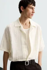 Camisa de calce relajado confeccionada en tejido de algodón color beige. Presenta un diseño de jacquard con líneas verticales desflecadas, cuello tipo bowling, manga corta y cierre frontal con botones.