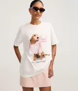 Remera blanca de algodón con cuello redondo y mangas cortas. Presenta una gran estampa central de un perro de raza cocker spaniel con un gran lazo rosa atado al cuello, y el texto "Do I look Pretty?" debajo.