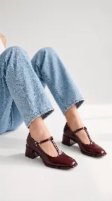 Zapatos de tacón bajo tipo Mary Jane con tira en T, confeccionados en charol color burdeos, con puntera cuadrada y cierre de hebilla lateral.