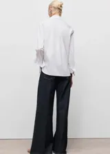 Camisa de corte recto en color blanco, con cuello camisero de solapas amplias, cierre frontal oculto con botones y mangas largas con puños abotonados.